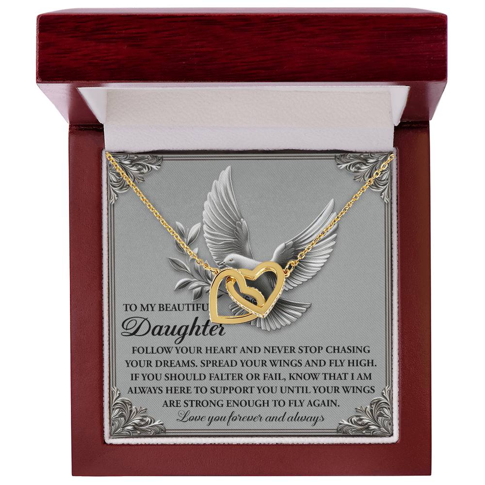 The Interlocking Hearts Necklace with cubic zirconia crystals features a gold heart with a bird design. It rests in an open wooden box, adorned inside with ornate corners and a bird image, plus an inspirational message to a daughter about following dreams and support.