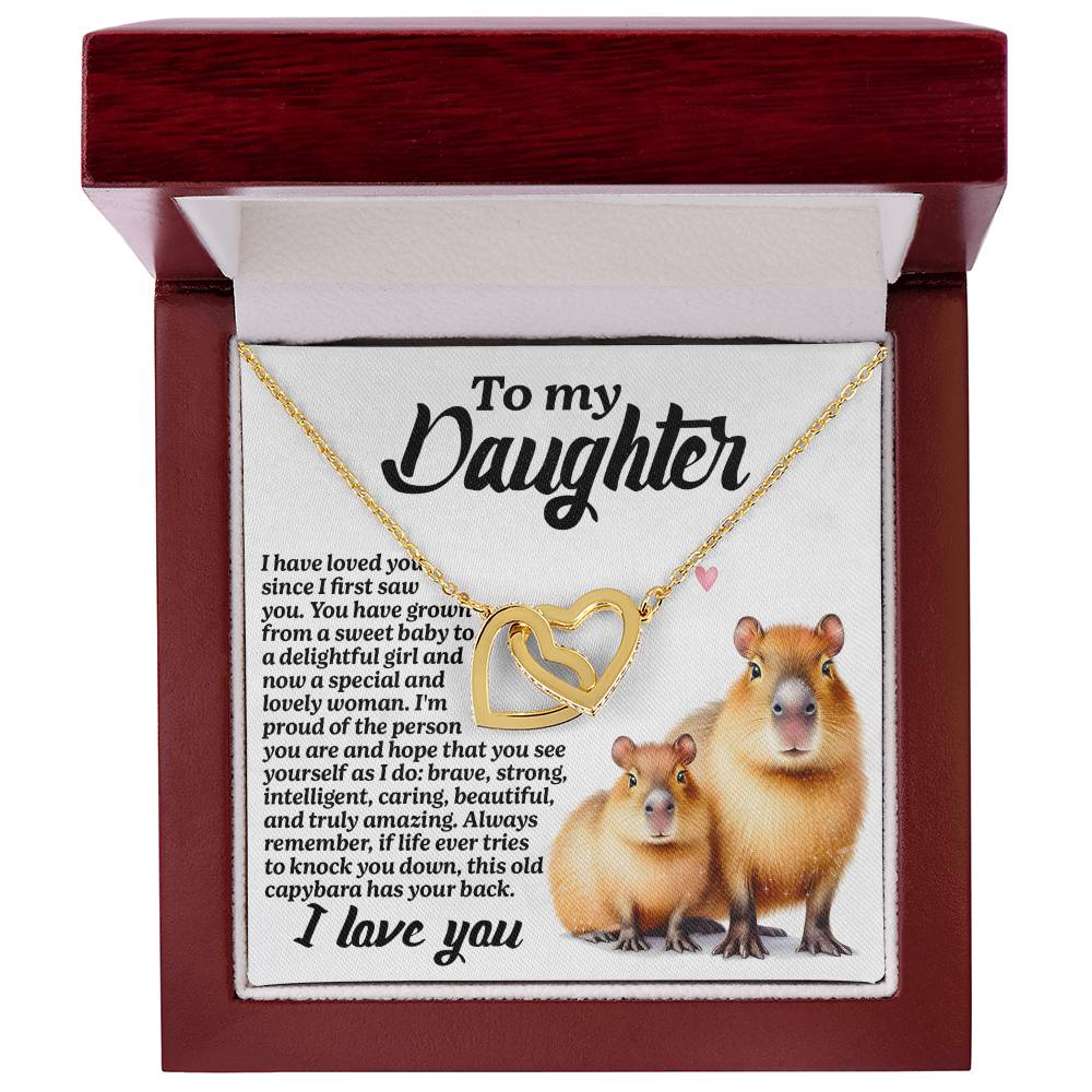 The Daughter-Sweet Baby Interlocking Hearts Necklace features 18k yellow gold finish, nestled in a wooden jewelry box with a white interior and sparkling cubic zirconia crystals. The lid displays a heartfelt message to a daughter and images of two capybaras at the bottom.