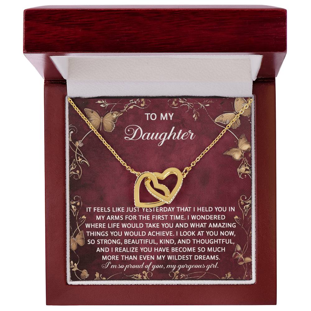 The Daughter-Wildest Dreams Interlocking Hearts Necklace, with a rose gold finish and cubic zirconia crystals, shines in a jewelry box. Its lid opens to reveal a heartfelt message for your daughter, expressing pride and admiration.