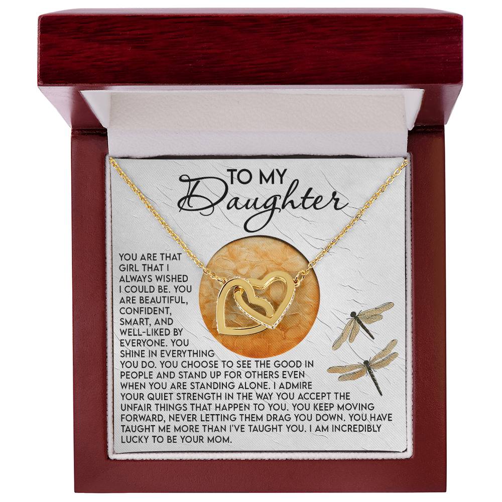 The Daughter-Quiet Strength - Interlocking Hearts Necklace adorned with cubic zirconia is nestled in a rose gold-finished jewelry box. Inside, a "To My Daughter" message conveys love and admiration, accompanied by two decorative dragonflies, while the box showcases a dark wood finish.