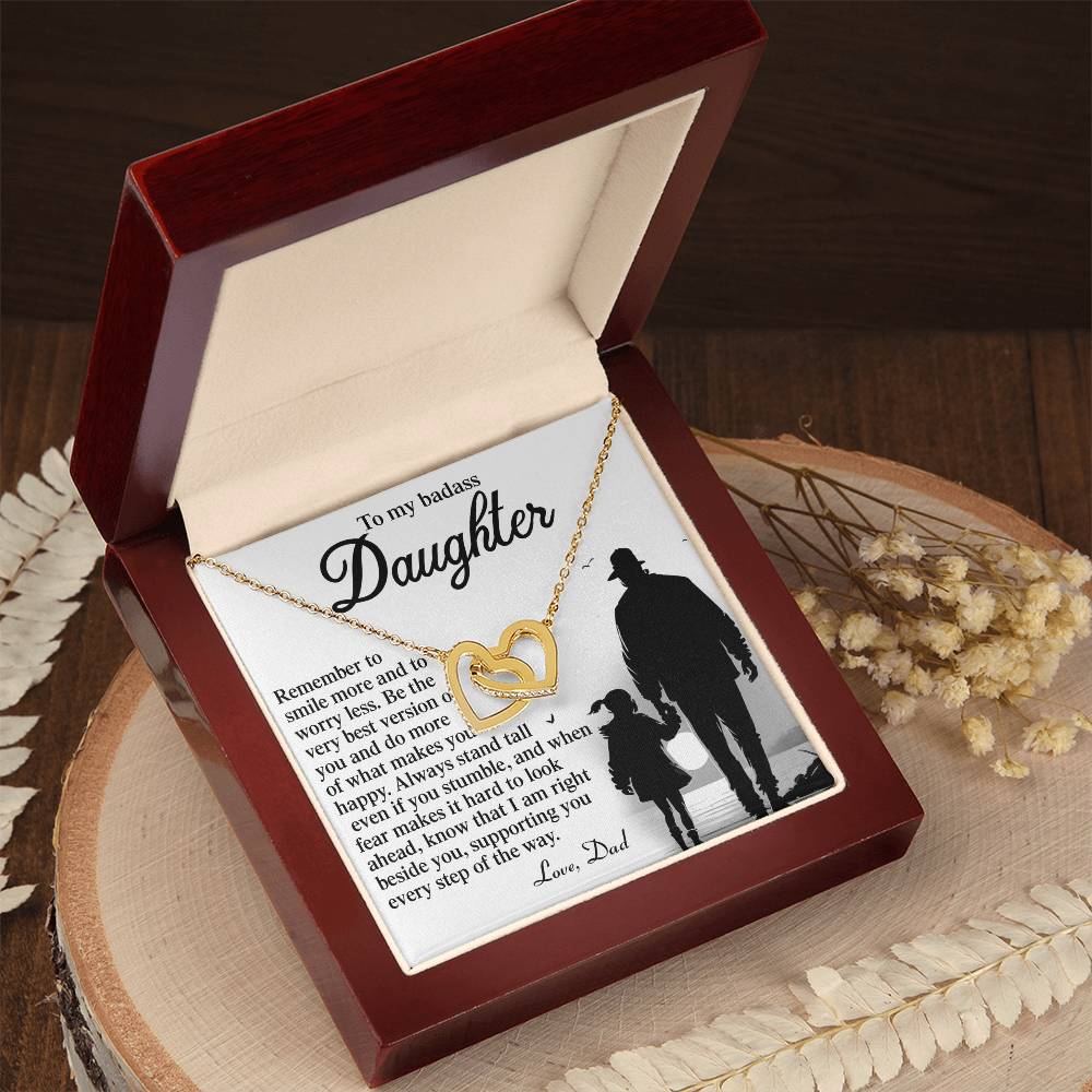 The Daughter-Smile More - Interlocking Hearts Necklace has a rose gold finish with an entwined smaller heart, presented in a wooden box with a heartfelt message from father to daughter on the lid. The box sits on a round wooden slab decorated with dried flowers.