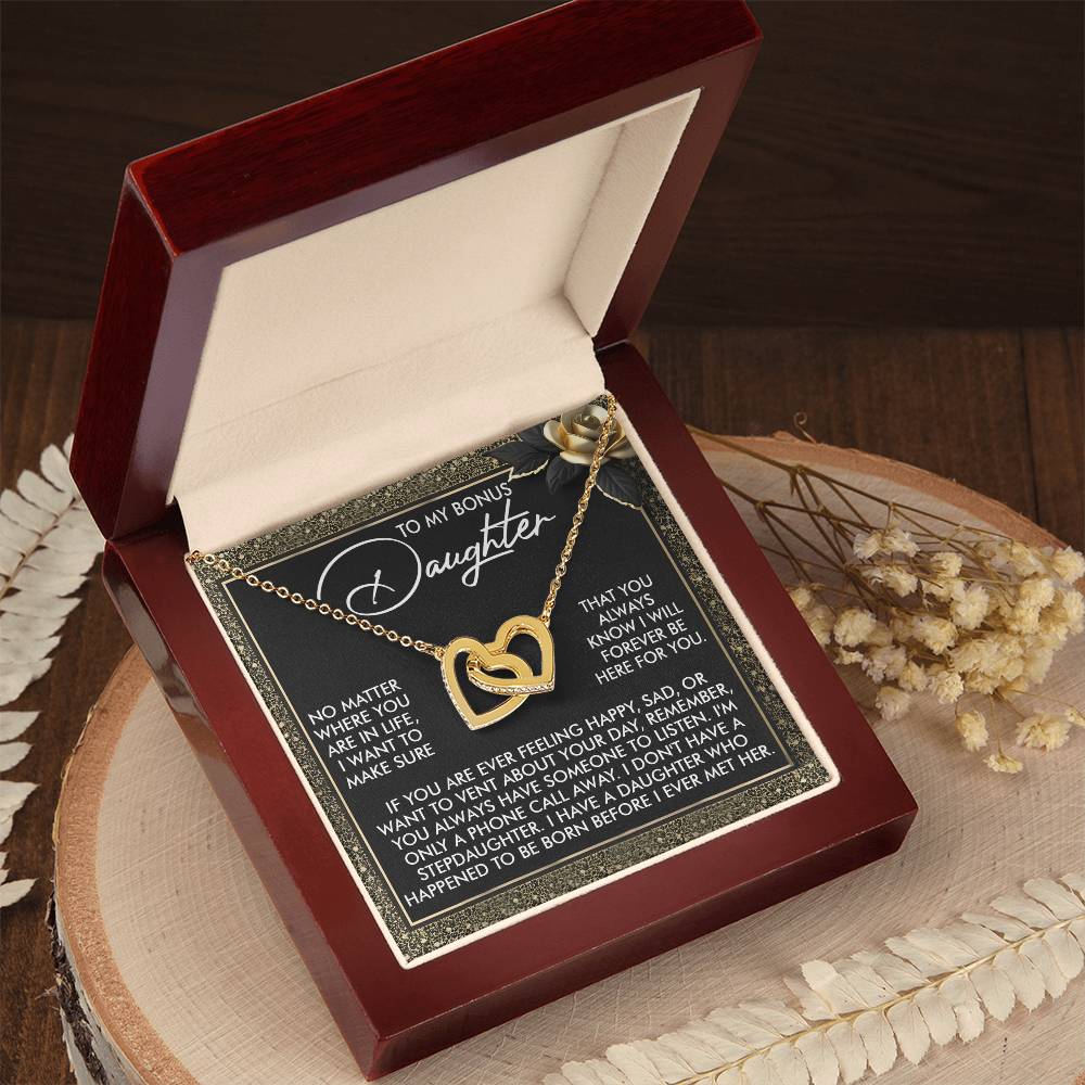 An open jewelry box displays the Daughter-Phone Call - Interlocking Hearts Necklace, embellished with cubic zirconia crystals. Bearing a heartfelt message for a "bonus daughter," it is crafted from surgical steel and rests on a wooden surface amidst decorative dried flowers.