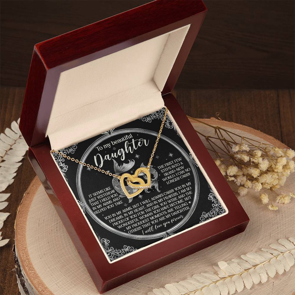 A gold "Daughter-In My Prayers" Interlocking Hearts necklace with cubic zirconia crystals is elegantly displayed in an open red velvet box, accompanied by a touching message on a circular card. Dried flowers and wooden slabs are arranged beneath the box.