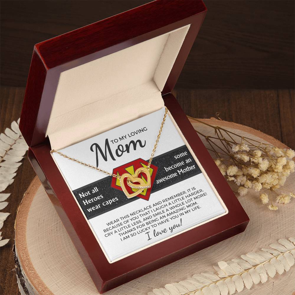 An open wooden box presents the Mom-Awesome Mother - Interlocking Hearts Necklace, embellished with cubic zirconia. The message reads, "To my loving Mom. Not all heroes wear capes. Some become an awesome mother." Dried flowers are gracefully arranged nearby on a wooden surface.