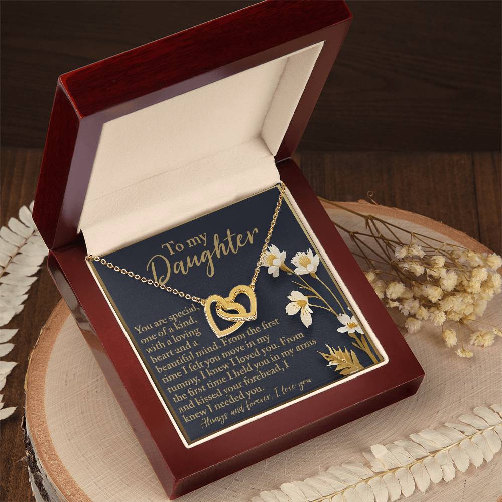 An open wooden box displays the "Daughter-One Kind - Interlocking Hearts Necklace" adorned with cubic zirconia, alongside a heartfelt message. The background features white daisies and decorative leaves on the wooden surface.
