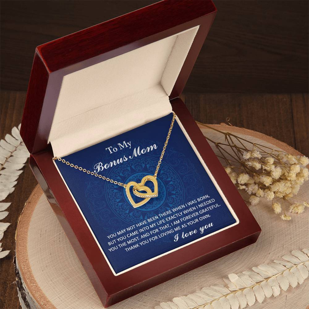 The Bonus Mom-I Needed You - Interlocking Hearts Necklace shines in rose gold with cubic zirconia crystals, housed in a wooden box featuring a touching message for a "Bonus Mom" on a blue background, accented by floral designs on wood.