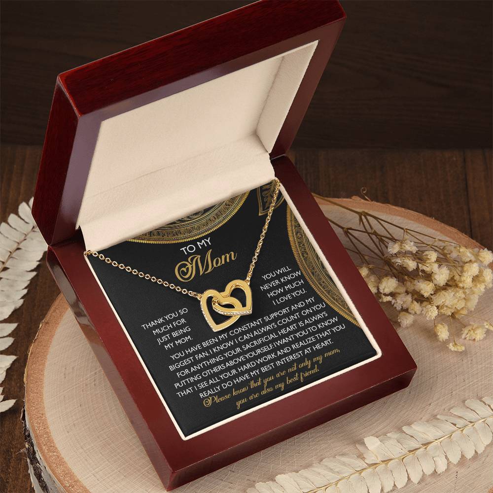 The Mom-Best Friend - Interlocking Hearts Necklace, with a rose gold finish, rests in a red velvet box featuring "Mom" on the background. Dried flowers and fern leaves accentuate the setting on a wooden surface, while cubic zirconia crystals gleam softly.