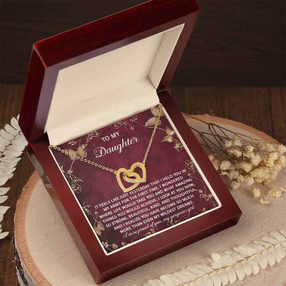 The Daughter-Wildest Dreams Interlocking Hearts Necklace, adorned with cubic zirconia crystals, is presented in an open red box with a heartfelt message and surrounded by dried flowers.