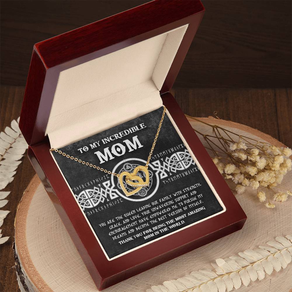 The "To Mom, The Queen 2 - Interlocking Hearts Necklace" is a personalized gold heart necklace set in a red velvet box adorned with cubic zirconia crystals and dried flowers, inscribed with "Mom," making it a truly special keepsake.