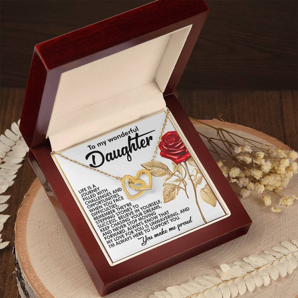 An open wooden box reveals a Daughter-Journey Challenges Interlocking Hearts Necklace in a rose gold finish. The lid displays a heartfelt message for a daughter with a red rose illustration, set against a wooden background with dried flowers to highlight the necklace's elegance.