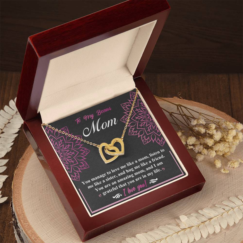 The Bonus Mom-An Amazing Mom Interlocking Hearts Necklace, adorned with cubic zirconia crystals, is presented in an open dark wooden box. A heartfelt, floral-patterned card for a "Bonus Mom" and dried flowers complement this elegant gift on a wooden surface.