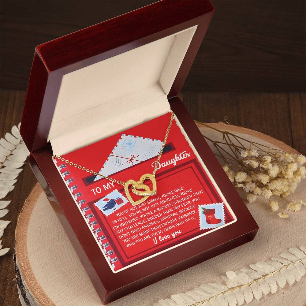 The red jewelry box is open, revealing the stunning Daughter-Part Of It - Interlocking Hearts Necklace inside, crafted with a rose gold finish and adorned with cubic zirconia crystals. A heartfelt note lies beneath the necklace, while dried flowers surround the box resting on a wooden surface.