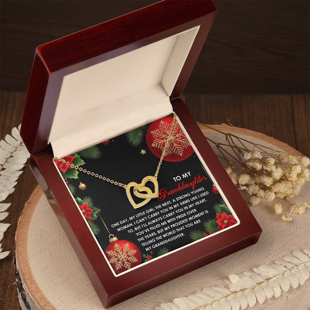 The Granddaughter-Proudest Moment - Interlocking Hearts Necklace, featuring a rose gold finish and elegant cubic zirconia crystals, is beautifully displayed in an open red jewelry box alongside a card. The card, decorated festively, holds a heartfelt message for a granddaughter, making this stunning piece the perfect gift.