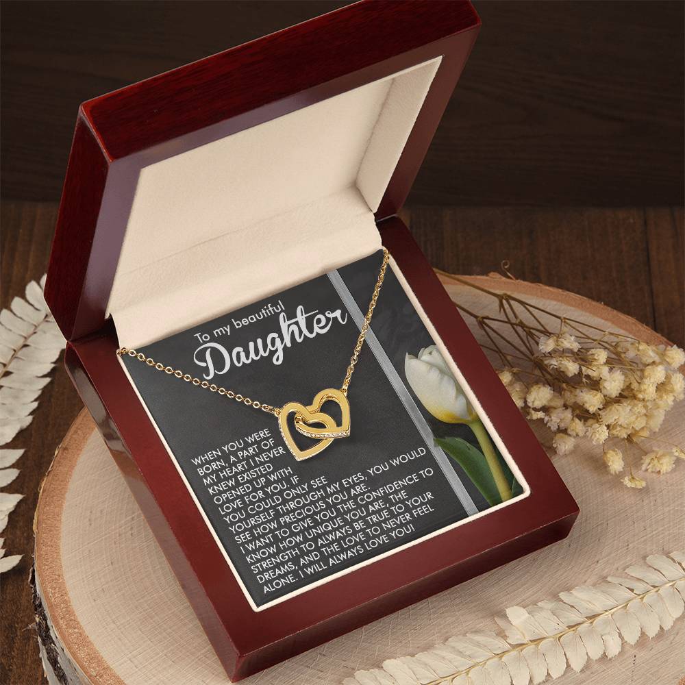 A Daughter-Heart Love Interlocking Hearts Necklace with a rose gold finish sits in an open wooden jewelry box. The lid shows a heartfelt message to a daughter alongside a yellow tulip card, complemented by white flowers arranged tastefully beside the box.