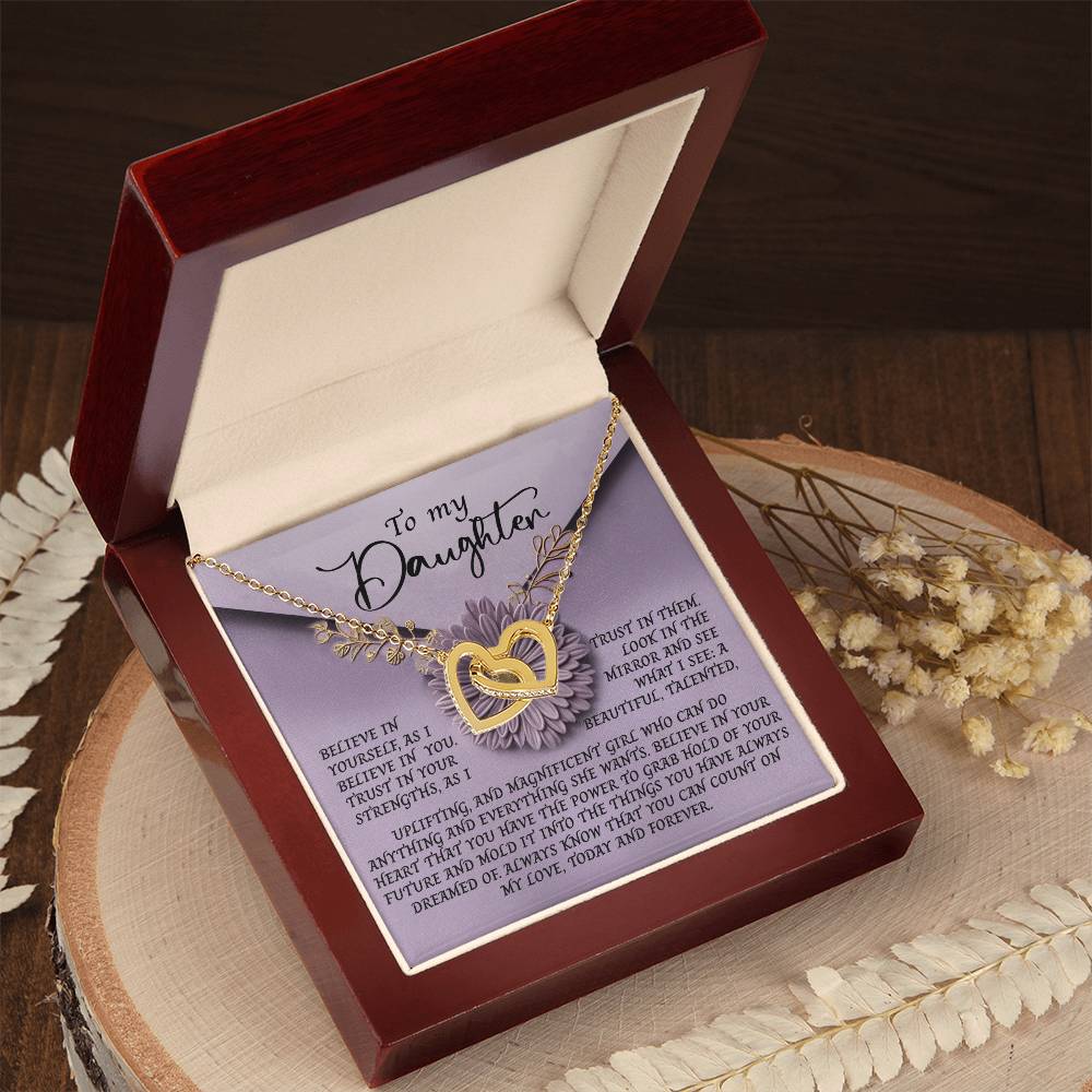 An open jewelry box reveals a Daughter-Magnificent Girl - Interlocking Hearts Necklace with cubic zirconia crystals. The inner lid displays a heartfelt message to a daughter with decorative borders, while the rose gold finish sparkles on a wooden surface amid dried flowers.