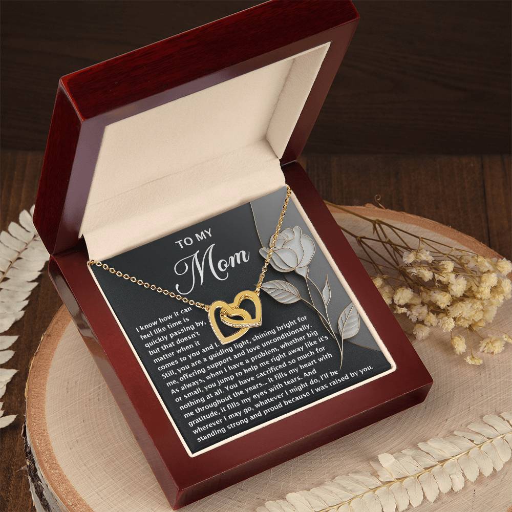 A Mom-Guiding Light - Interlocking Hearts Necklace with a rose gold finish rests in a dark red jewelry box. A "To My Mom" card, featuring a rose design and heartfelt message, stands behind. The box sits on a wooden surface adorned with decorative dried flowers for elegance.