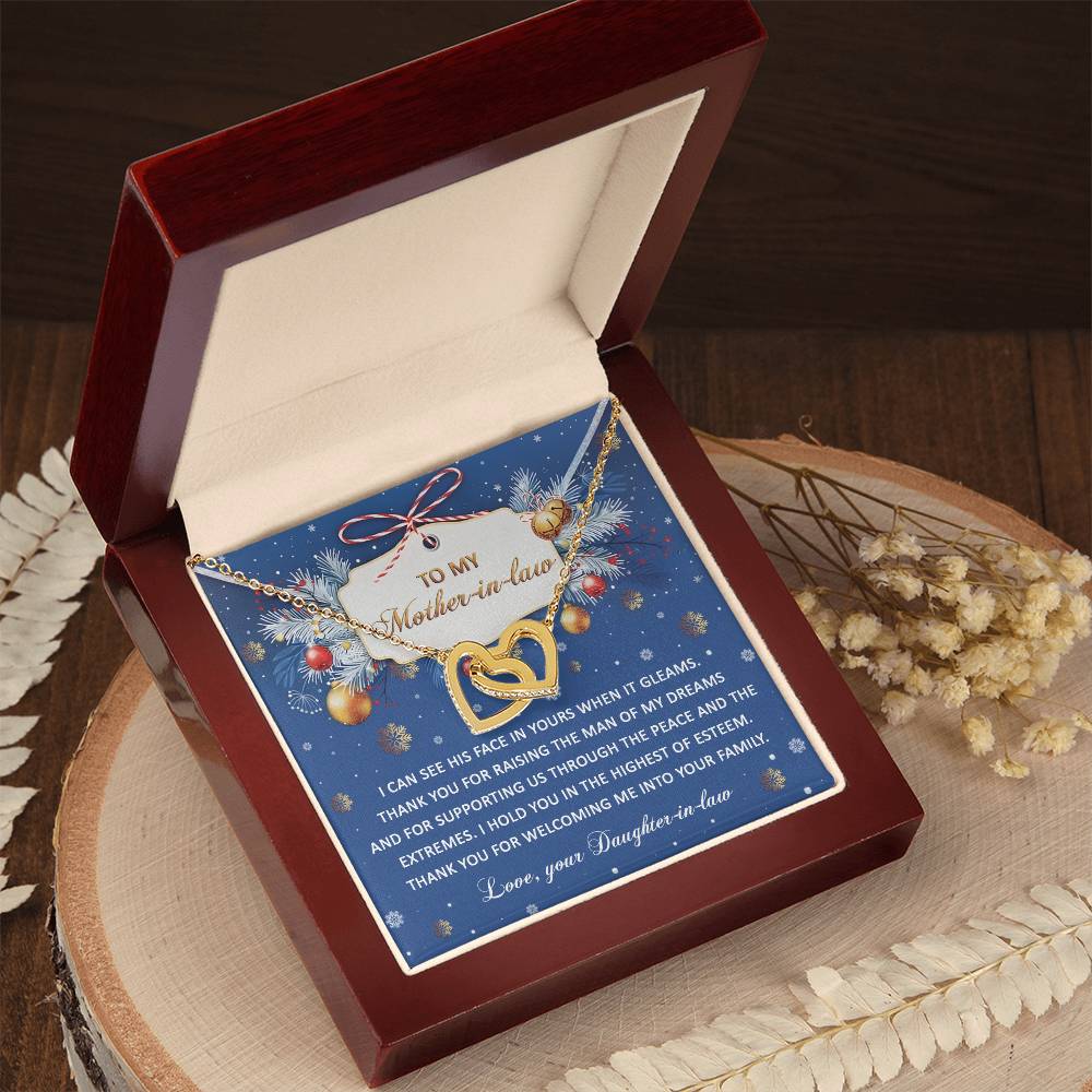 The "Mother-in-law-I Hold You - Interlocking Hearts Necklace" with a rose gold finish and adorned with cubic zirconia crystals is elegantly displayed in a box labeled "To My Mother-In-Law" on a wooden surface accented by decorative foliage.