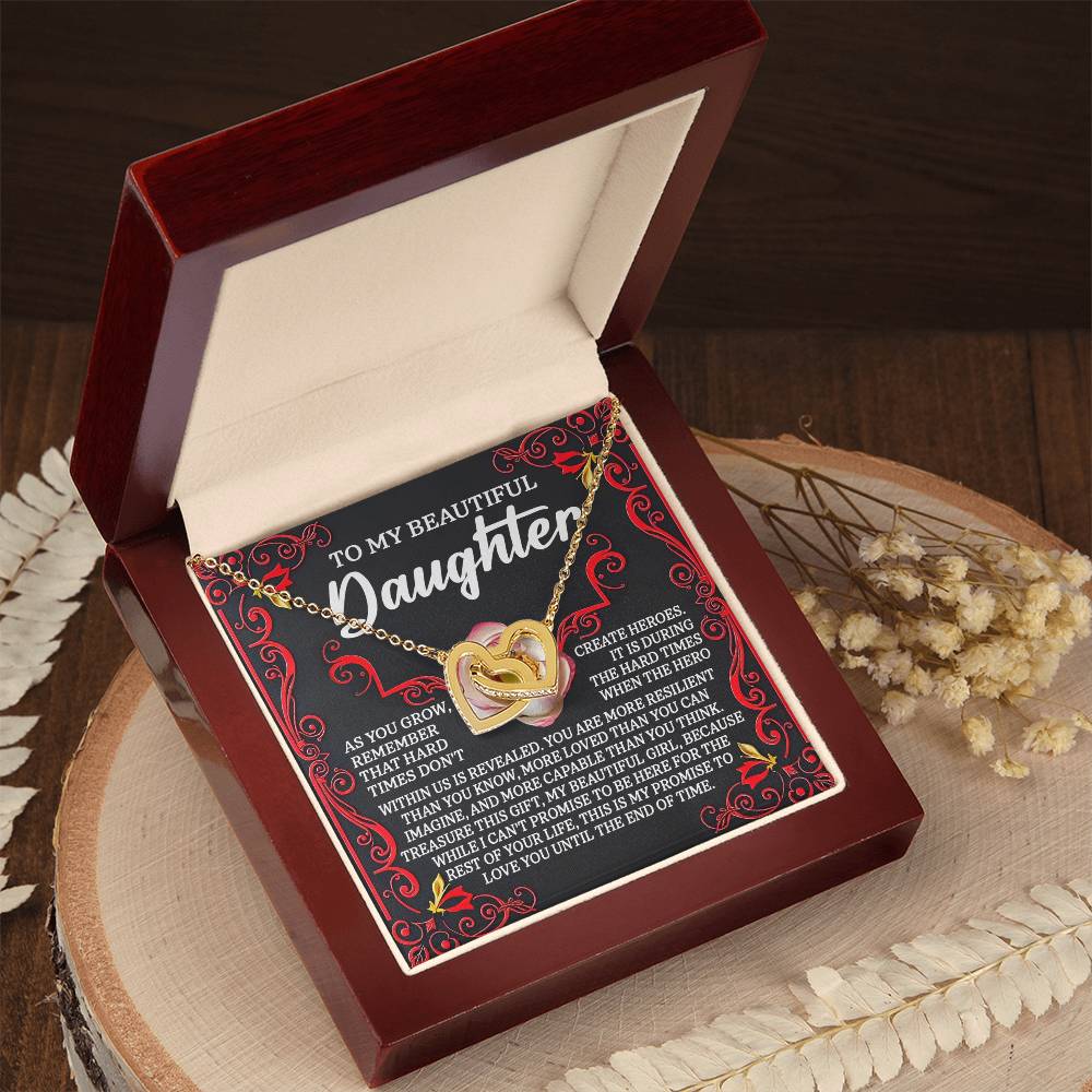 An open jewelry box presents the "Daughter-Hard Times" Interlocking Hearts Necklace, featuring a gold heart-shaped pendant with cubic zirconia crystals atop a heartfelt message for a daughter, surrounded by dried flowers on a wooden surface.