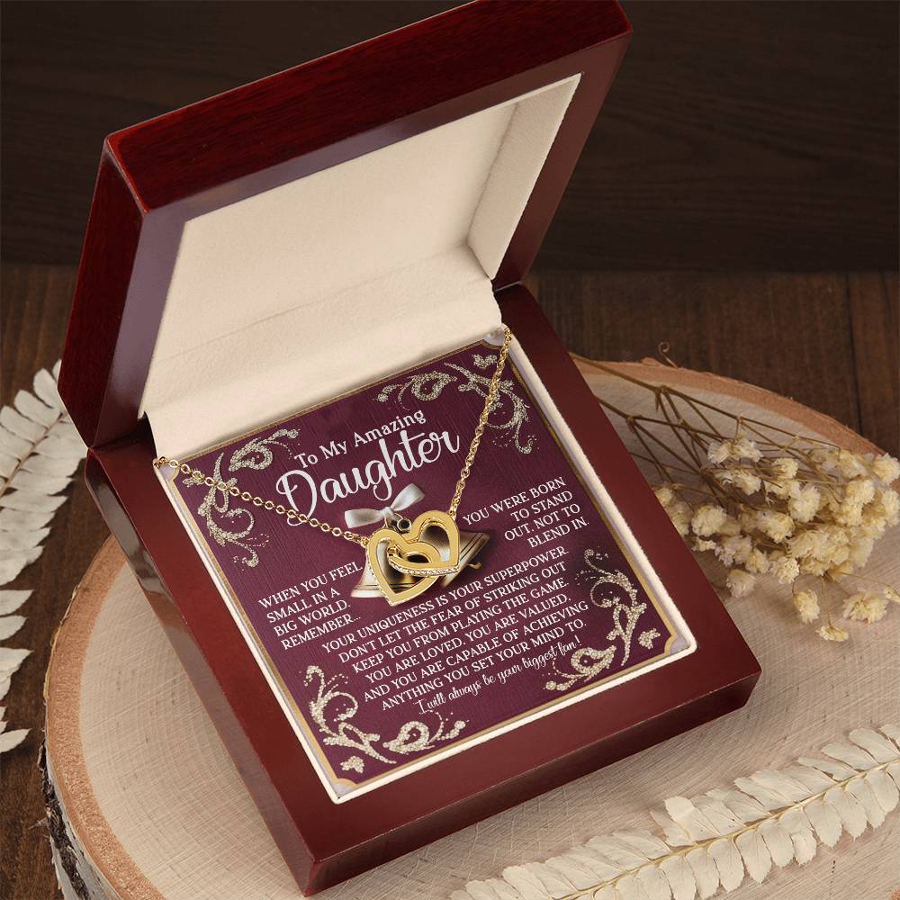 An open wooden jewelry box contains the Daughter-Stand Out - Interlocking Hearts Necklace, featuring cubic zirconia crystals and a gold heart design. The lid is inscribed for a daughter, celebrating her strength and love, with dried flowers beside it on the wooden surface.