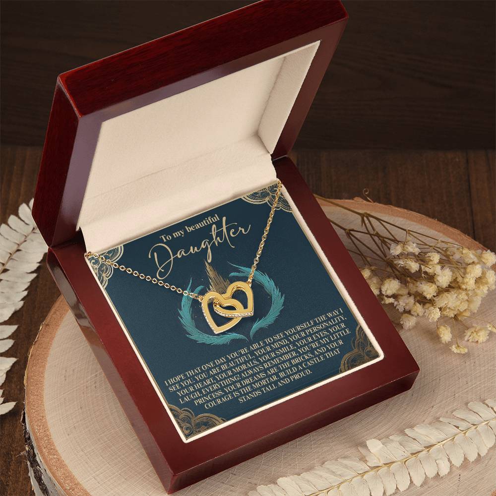 The Daughter-Little Princess Interlocking Hearts Necklace features a rose gold finish with sparkling cubic zirconia crystals, elegantly displayed in an open wooden box on a round surface adorned with dried flowers. Inside, a card reads, "To my beautiful daughter.