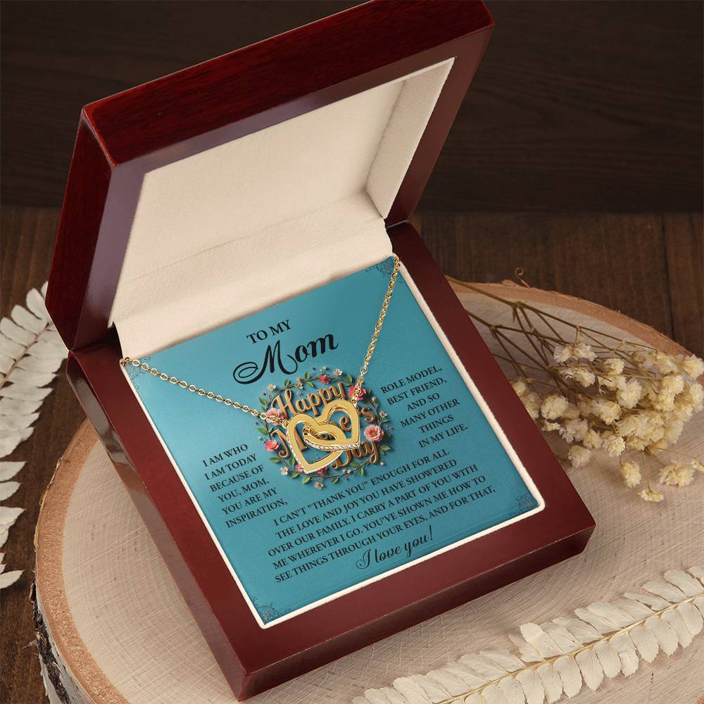The "Mom-Our Family" Interlocking Hearts Necklace in rose gold features a heart pendant with "Mom" and sparkles with cubic zirconia crystals. It comes in an elegant gift box that opens to reveal a heartfelt poem, resting on a round wooden surface amid decorative dried flowers.