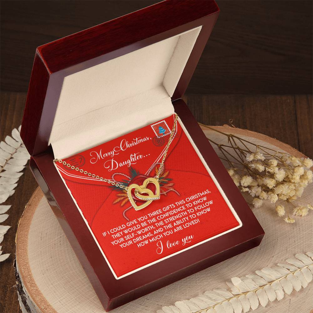 An open wooden box reveals the Daughter-This Christmas - Interlocking Hearts Necklace, featuring a shimmering rose gold finish. It is elegantly nestled on a red and white card with a heartfelt Christmas message for a daughter. The card is gracefully resting on a round wooden surface adorned with dried flowers.