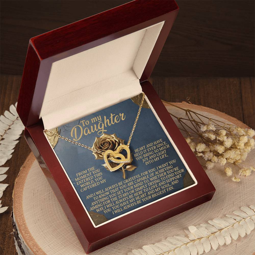 The Daughter-Captured Heart necklace, featuring interlocking hearts with a rose gold finish and a rose, shines in a red velvet box inscribed with "To my daughter" and floral motifs. The box sits on wood surrounded by dried flowers, enhancing its delicate charm.