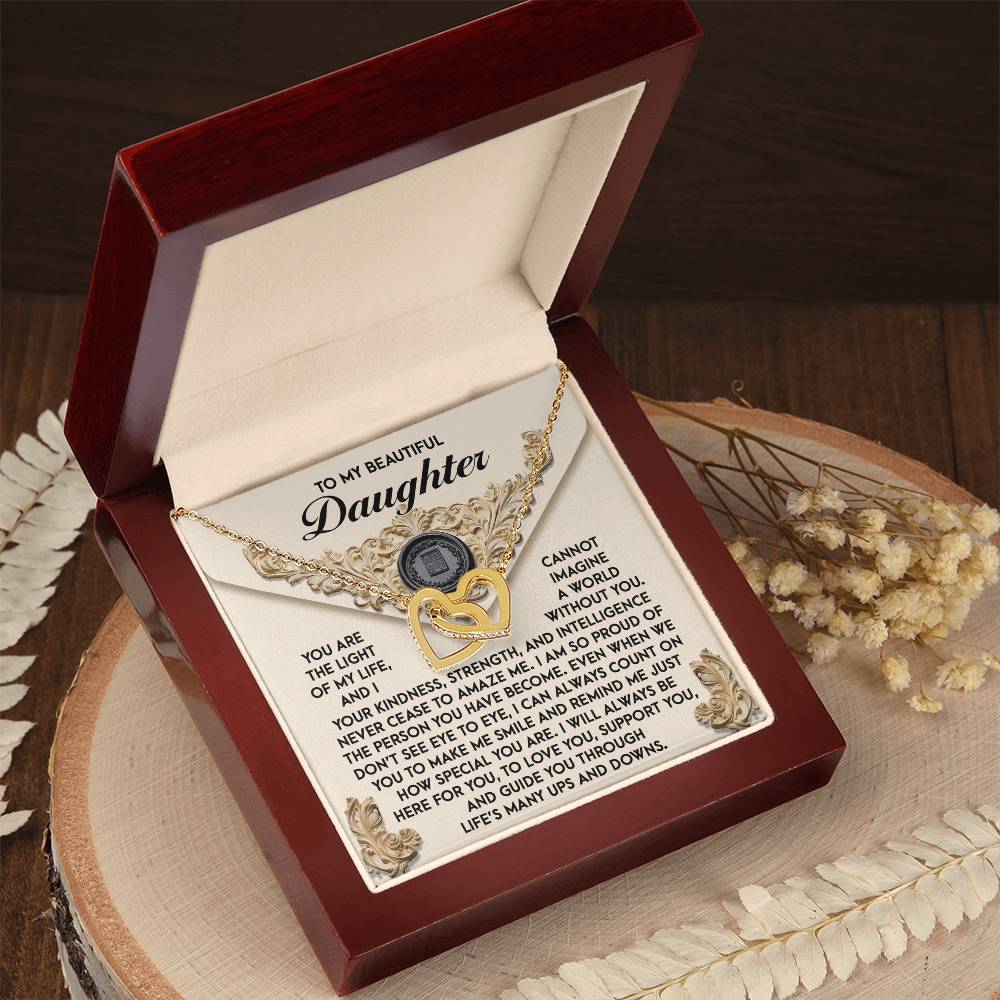 A wooden box with an open lid displays the "Daughter-Amaze Me" Interlocking Hearts Necklace, adorned with cubic zirconia crystals. Inside is a sentimental message for a daughter. White flowers accentuate the rose gold finish, adding elegance and warmth to the scene.