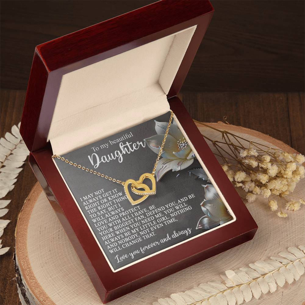 The Daughter-Love Protect Necklace, featuring interlocking hearts and a rose gold finish, is elegantly displayed in an open wooden gift box. The box is lined with a floral design and a heartfelt message for a daughter, complemented by dried flowers on the wooden surface.
