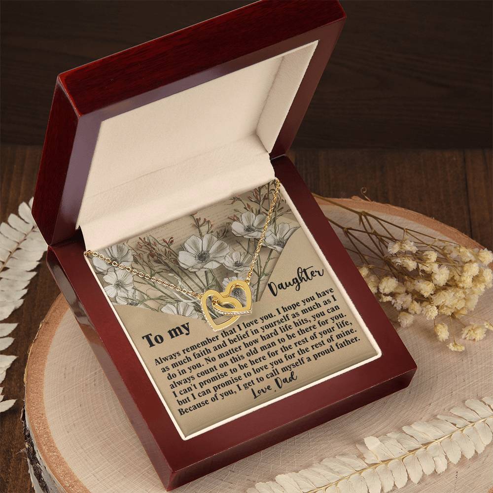 Inside an open wooden box, the Daughter-Proud Father - Interlocking Hearts Necklace sparkles with cubic zirconia. The rose gold finish complements a heartfelt message from a father to his daughter printed inside the lid against dried flowers, expressing love and pride.