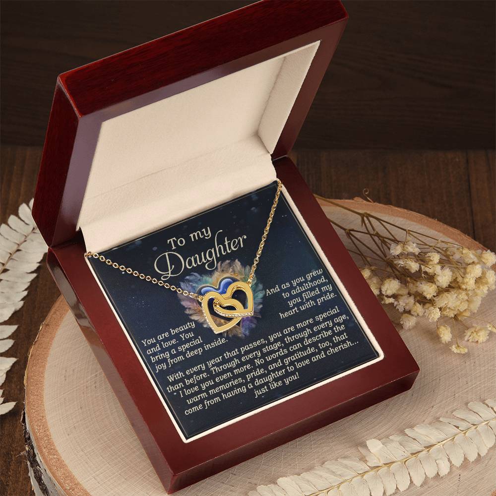 The Daughter-Like You Interlocking Hearts Necklace, adorned with cubic zirconia crystals, glistens in a red velvet-lined wooden box. Inside the lid is a heartfelt "To my Daughter" message. The box is placed on wood and surrounded by dried flowers.