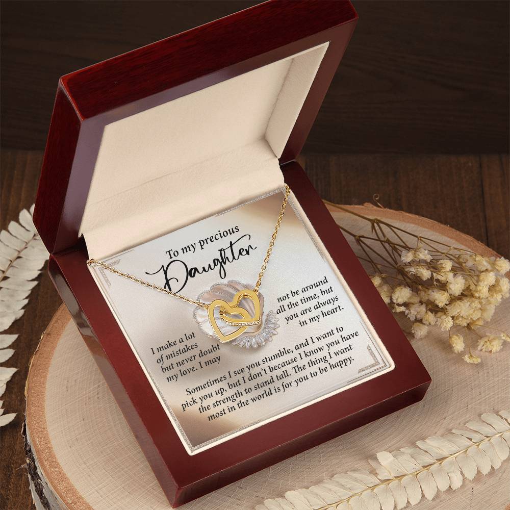 The "Daughter-Be Happy - Interlocking Hearts Necklace," featuring a rose gold finish with cubic zirconia crystals, rests in a red jewelry box engraved with a heartfelt message. The box sits elegantly on a wooden surface, surrounded by dried flowers and leaves.