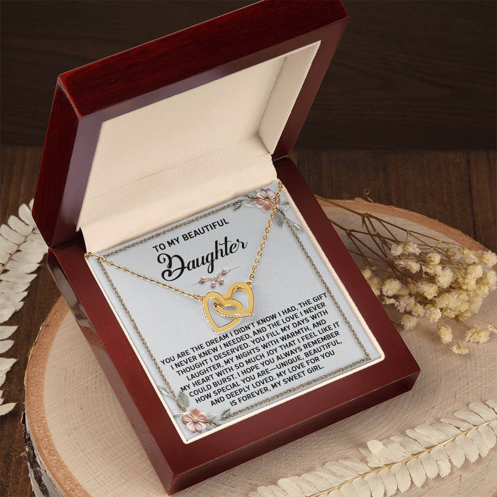 An open box displays the Daughter-My Sweet Girl Interlocking Hearts Necklace, rose gold with sparkling cubic zirconia, accompanied by a heartfelt message to a daughter, elegantly set on a wooden surface adorned with decorative flowers.
