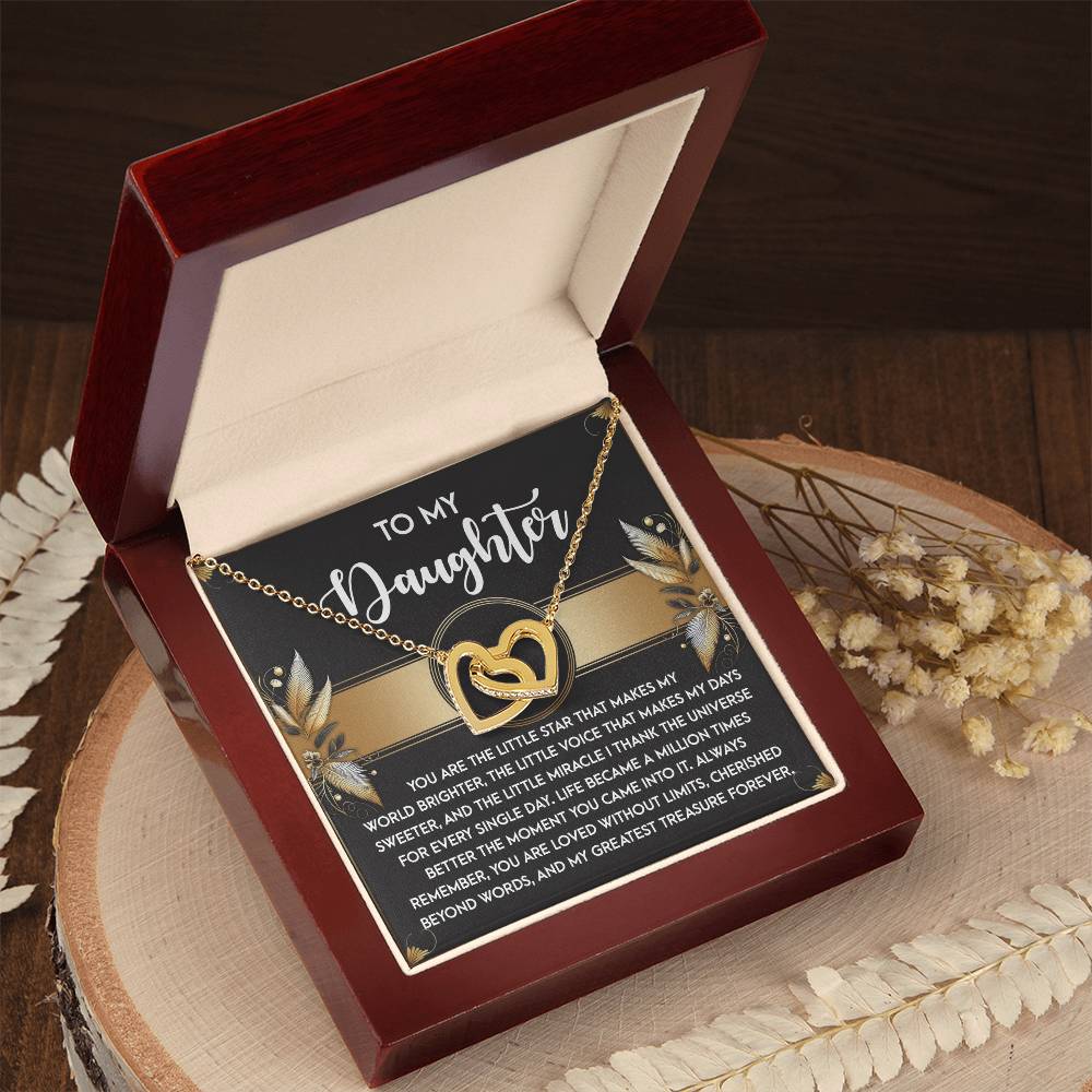The Daughter-Greatest Treasure Interlocking Hearts Necklace, with a rose gold finish and shimmering cubic zirconia crystals, rests in a red box with a heartfelt message inside. It's nestled on a wooden surface adorned with delicate dried flowers.