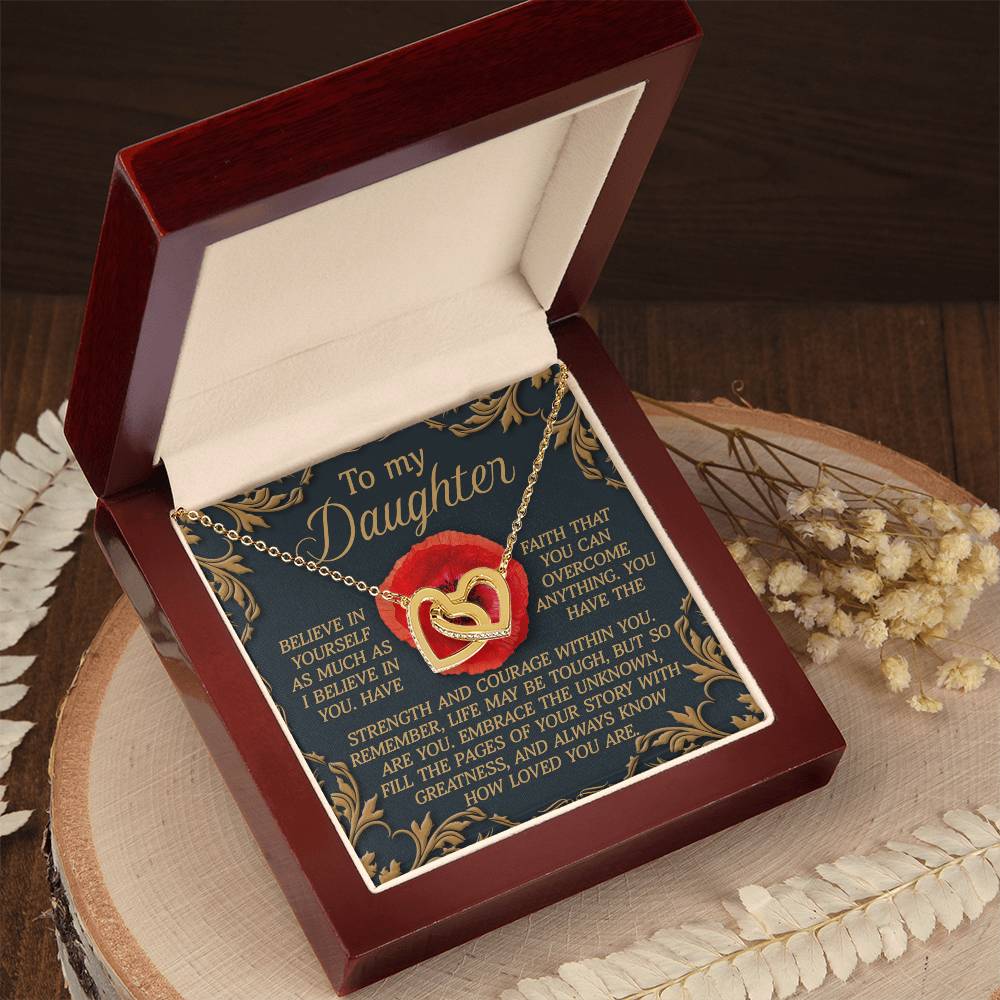 An open red and beige jewelry box features the "Daughter-With Greatness" Interlocking Hearts Necklace, embellished with cubic zirconia. Inside the lid, a touching message to a daughter is framed by an elegant border. The box elegantly rests on wood, enhanced by dried flowers nearby.