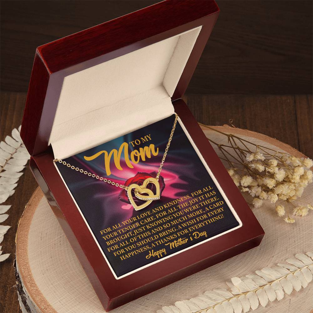 The Mom-Tender Care - Interlocking Hearts Necklace, featuring sparkling cubic zirconia crystals, comes in a red velvet box with a "To My Mom" message card ending with "Happy Mother's Day." The box is placed on a wooden surface, surrounded by dried flowers.