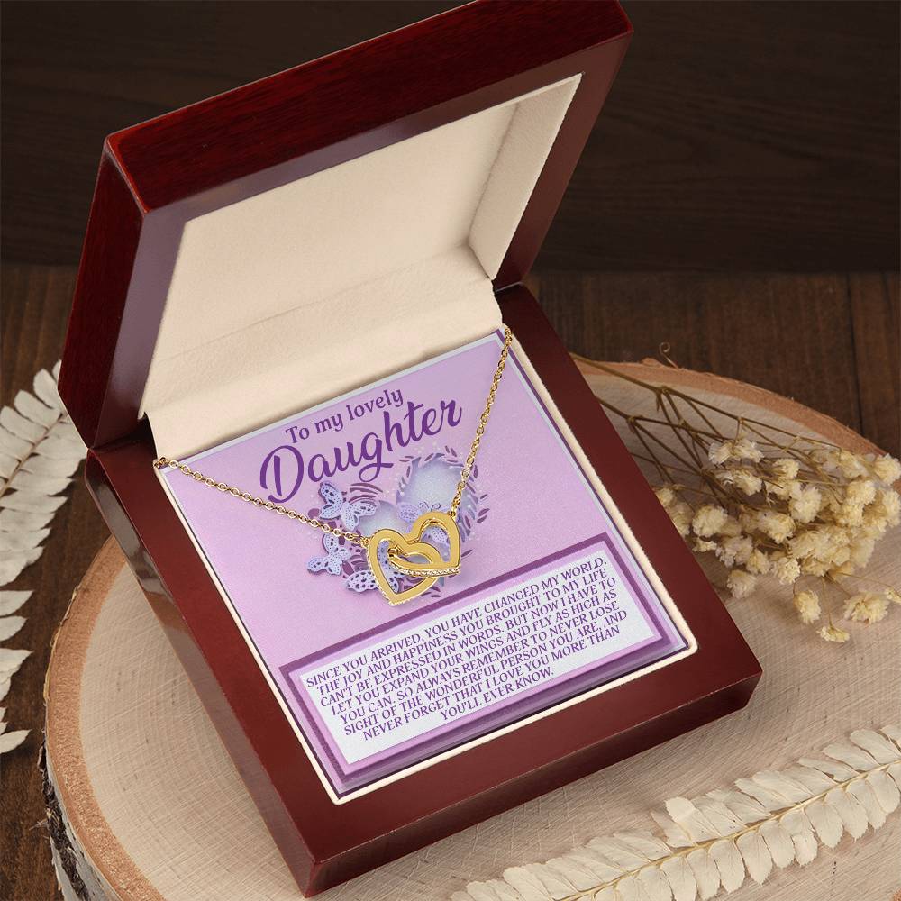 The Daughter-Expand Wings - Interlocking Hearts Necklace, with a rose gold finish, elegantly rests in an open red jewelry box. The lid reads "To my lovely Daughter," and the base features a heartfelt message. Decorative dried flowers add charm to the display.