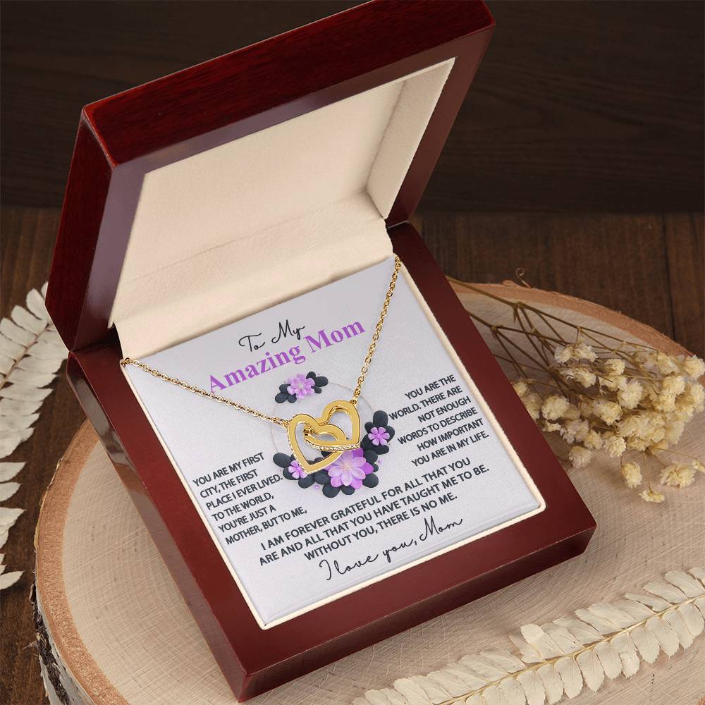 An open wooden box reveals the Mom-Forever Grateful - Interlocking Hearts Necklace, featuring a rose gold finish with cubic zirconia and purple flowers. The lid displays a heartfelt message for "My Amazing Mom," surrounded by small purple flowers, resting on a wooden surface with dried leaves.