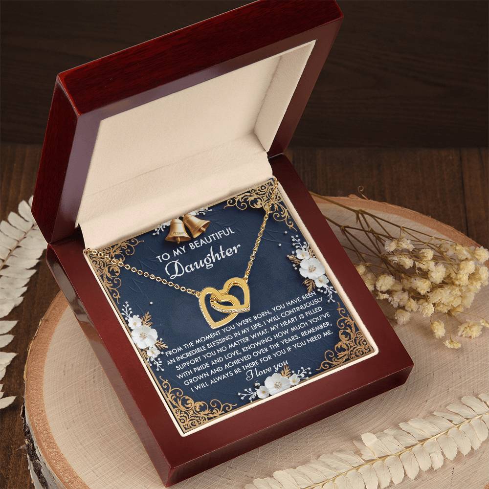 Open jewelry box featuring the "Daughter-If You - Interlocking Hearts Necklace" with cubic zirconia crystals. Inside is a floral card addressed to "Daughter," expressing love and pride, placed on a wooden surface surrounded by white flowers.