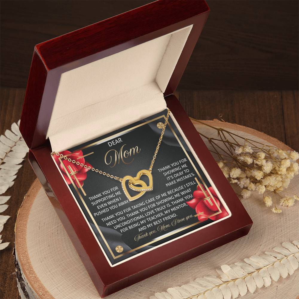 The "Mom-Best Friend 2 - Interlocking Hearts Necklace" in gold with a rose gold finish is showcased in an open red velvet box. Inside is a heartfelt card for "Mom," adorned with red roses, on a wooden surface surrounded by dried flowers.
