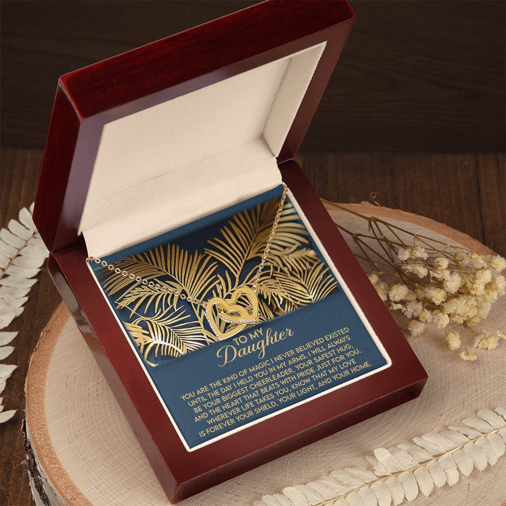 A Daughter-Your Home Interlocking Hearts Necklace with a rose gold finish is nestled in a red and cream jewelry box. The box, showcasing "To My Daughter" on a dark background with leaf patterns, rests on wood near dried flowers.