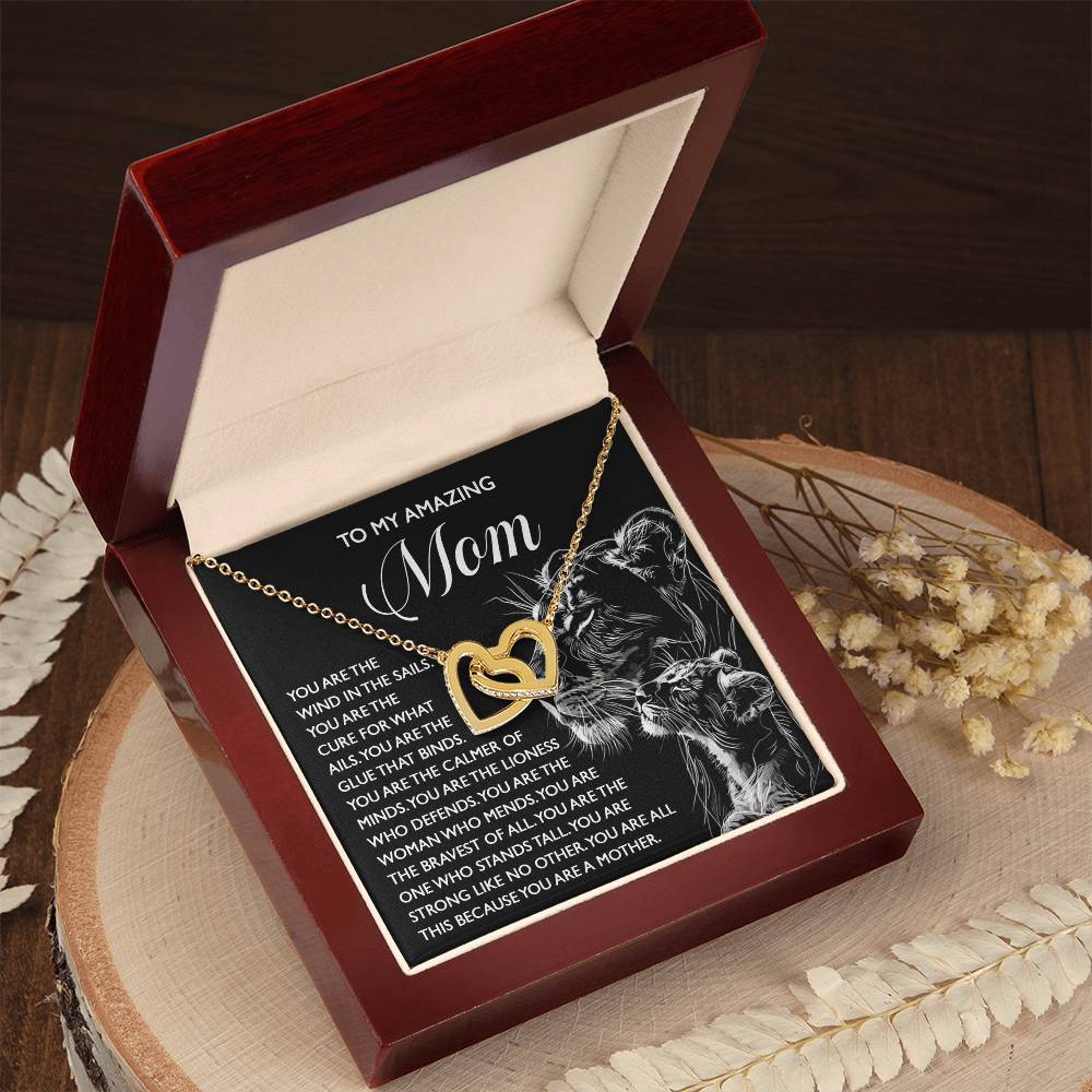 An open jewelry box reveals a stunning Mom-The Cure - Interlocking Hearts Necklace in rose gold finish, resting on an engraved "To My Amazing Mom" message. Nearby, decorative dried flowers lie gracefully on the wooden surface.