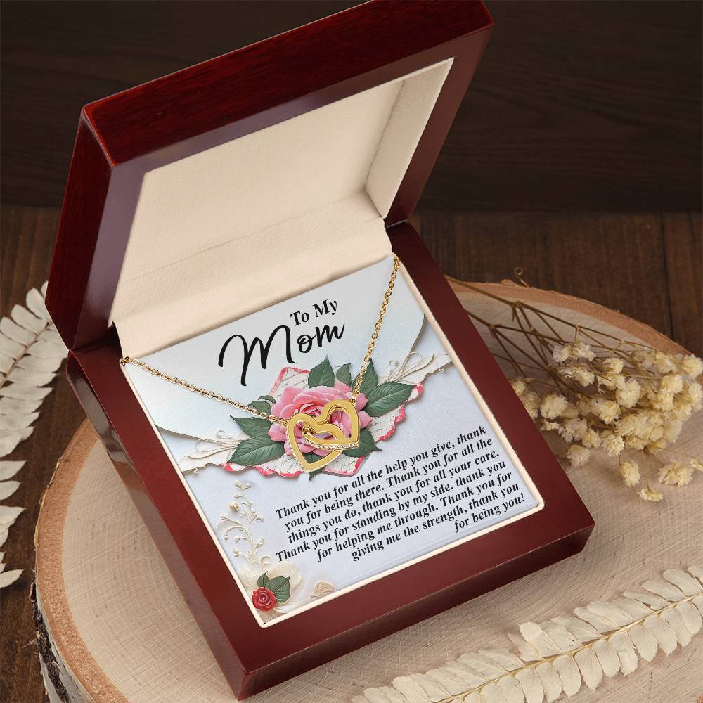 The Mom-Being You - Interlocking Hearts Necklace, with a rose gold finish, is elegantly nested in a red gift box. A heartfelt message for mom embellishes the lid, decorated with pink and yellow roses. The box sits on wood amidst dried flowers and sparkling cubic zirconia crystals.