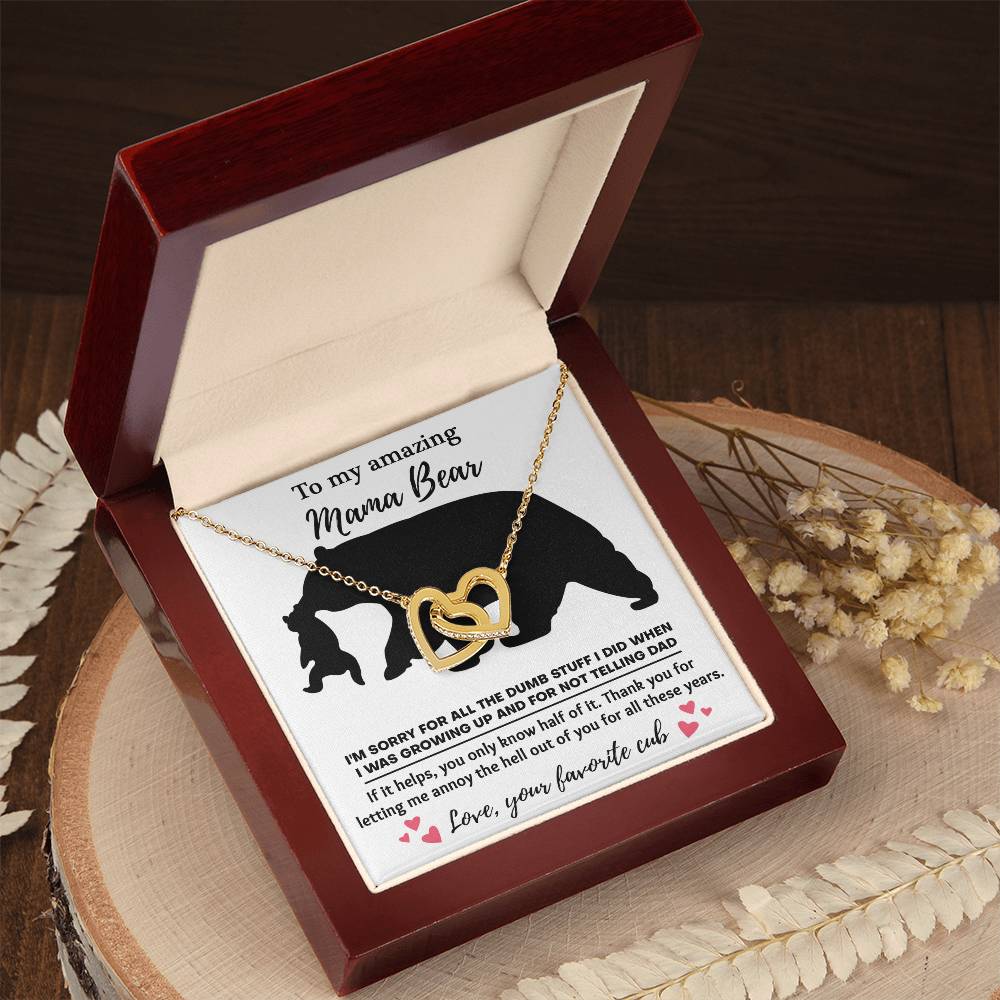 The "To Mom, Not Telling Dad - Interlocking Hearts Necklace" is a gold necklace with cubic zirconia, presented in a wooden box. It includes a heartfelt note featuring a bear silhouette and an apology message, making it perfect for the Mama Bear in your life.