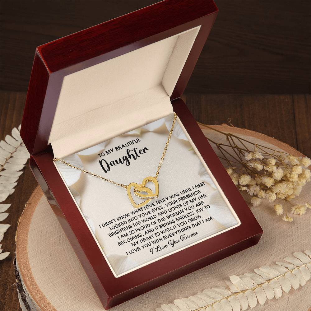 The Daughter-Into Your Eyes Interlocking Hearts Necklace features a gold heart pendant in a wooden box lined with cubic zirconia. The inner lid bears a heartfelt message for a daughter, and elegant white flowers complete this perfect gift.