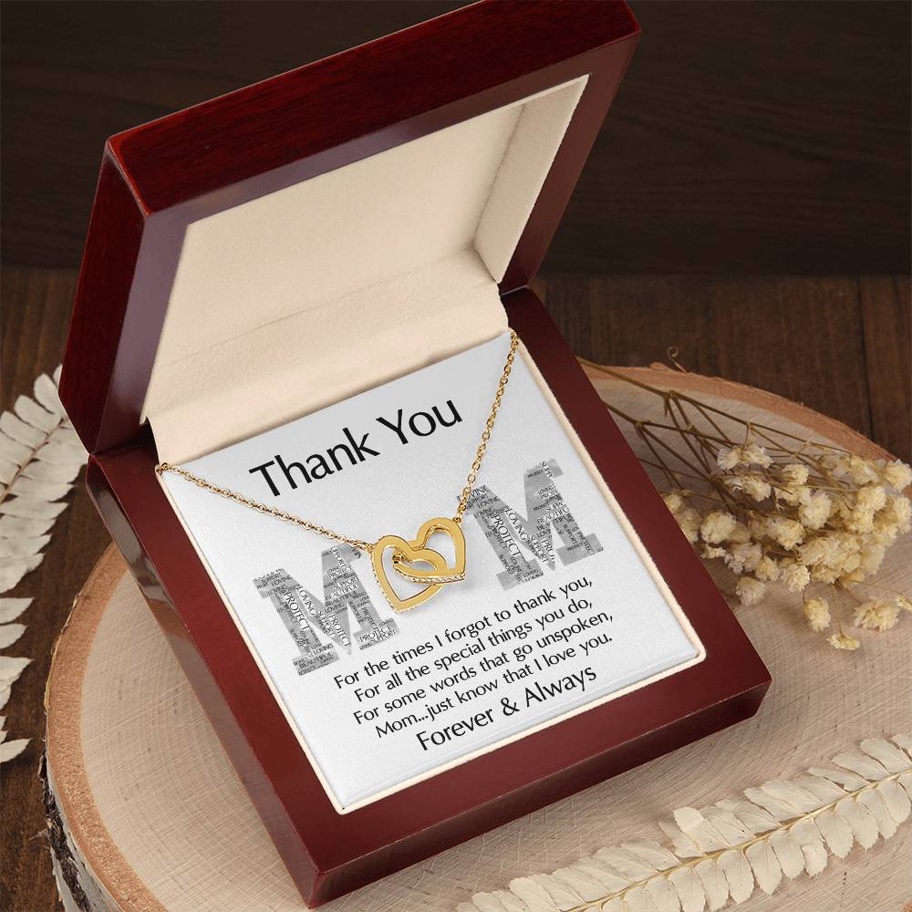 The "To Mom, Thank You - Interlocking Hearts Necklace," adorned with cubic zirconia crystals, comes in a wooden box with a "Thank You Mom" card. It rests on wood and is accompanied by dried flowers, embodying love and gratitude.