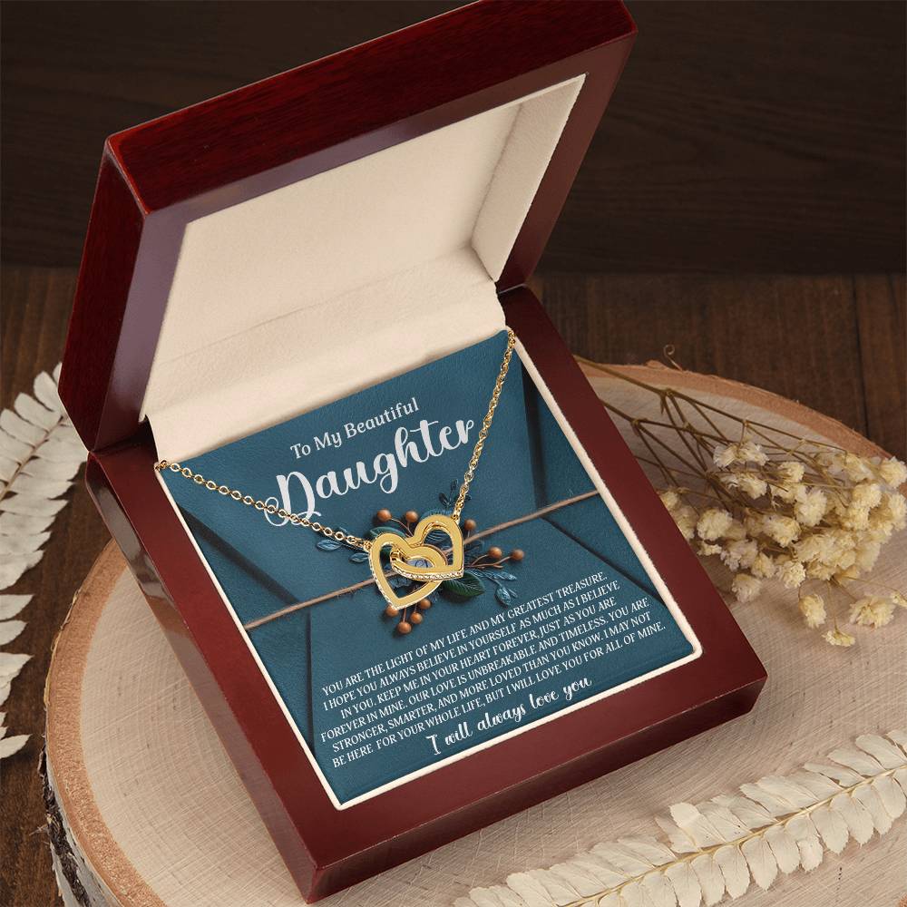 A Daughter-All Of Mine Interlocking Hearts necklace with a rose gold finish sits in an open velvet-lined jewelry box, the lid displaying a heartfelt message for a daughter. Dried flowers are nearby on the wooden surface, and cubic zirconia crystals provide a touch of sparkle.