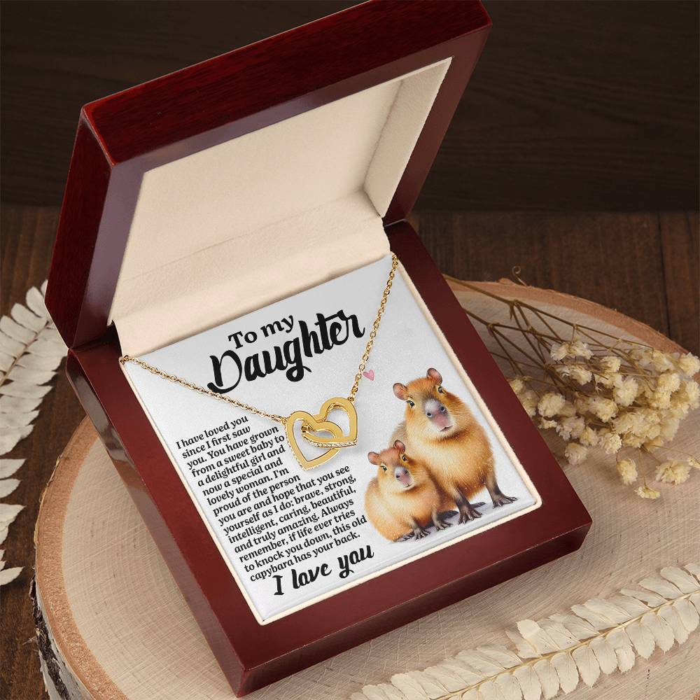 On a wooden surface, an open jewelry box reveals the Daughter-Sweet Baby - Interlocking Hearts Necklace with cubic zirconia crystals. Inside, a heart-shaped pendant shows two hamster-like animals, and the lid displays a heartfelt "To my Daughter" message, surrounded by scattered dried flowers.
