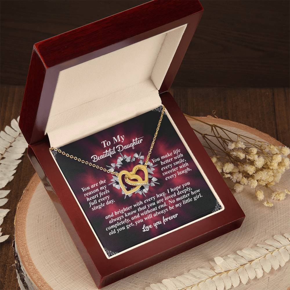 The Daughter-Every Hug - Interlocking Hearts Necklace, with a rose gold finish and adorned with cubic zirconia, is presented in an open red velvet-lined box. A heartfelt letter to a daughter accompanies it, while decorative white leaves and dried flowers grace the wooden surface nearby.