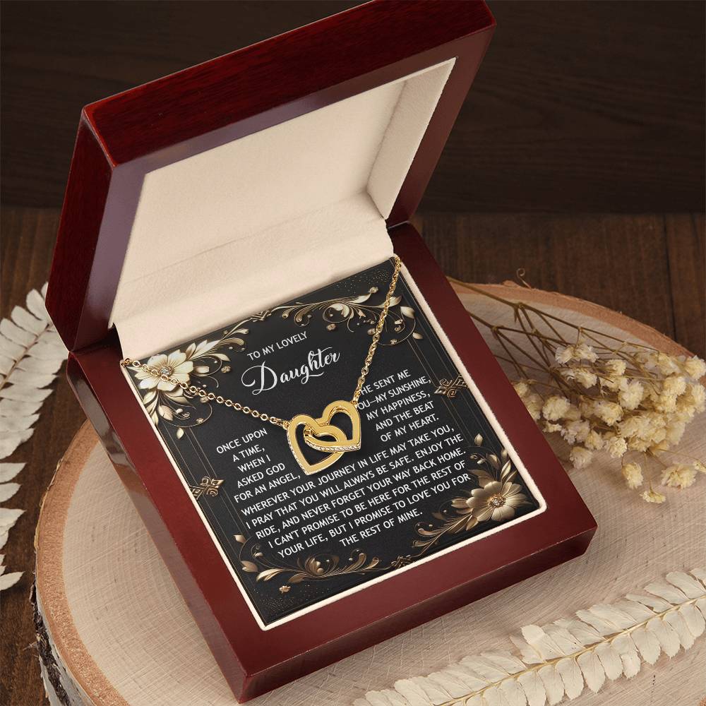The Interlocking Hearts Necklace, named "Daughter-Be Safe," glows with an 18k gold finish and cubic zirconia accents inside a red jewelry box. A heartfelt message lies within, and the elegant box is set on a wooden surface among dried flowers.